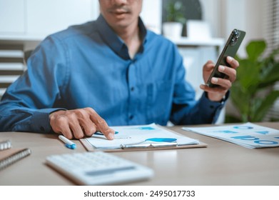 chart, document, graph, invoice, job, paperwork, planning, profit, report, smartphone. A man is sitting at a desk with a cell phone in his hand. He is looking at a piece of paper with a graph on it.
