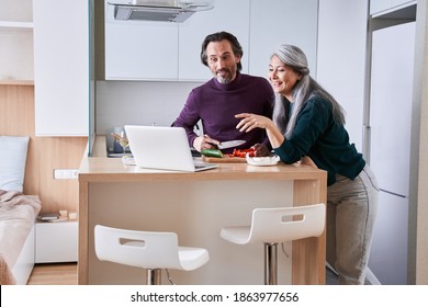 Chatting With Friends During Pandemic. Senior Loving Couple Calling To The Friends Via Skype While Preparing Dinner At The Modern Kitchen. Stock Photo