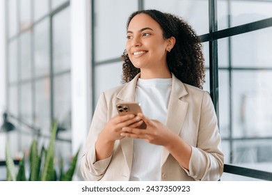 Chatting online. Positive multiracial stylish young woman with curly hair, stands in a modern office, messaging by smartphone with friends or clients online, checking news, looks away, smiling - Powered by Shutterstock