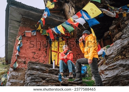 Chatting smiling Backpackers Couple tea break at small sacred Buddhist monastery decorated multicolored Tibetan prayer flags with mantras. Climbing Mera peak route in Makalu Barun National Park, Nepal
