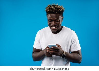 Chatting With You. Portrait Of Young Man In White T-shirt Using Mobile Phone And Smiling. Isolated On Blue Background