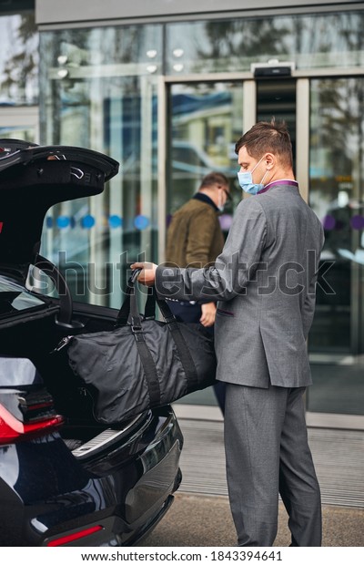 Chauffeur in a disposable face mask taking his\
client bag out of the car\
trunk