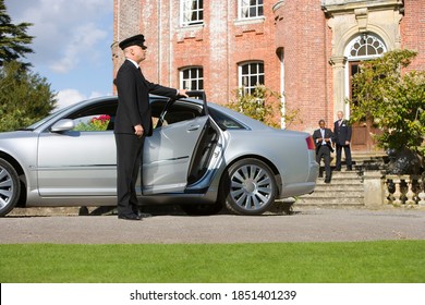 Chauffeur Holding The Car Door Open For Two Senior Businessmen In The Background In Front Of A Huge Manor House