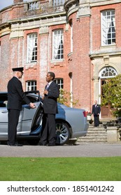 Chauffeur Holding The Car Door Open For A Senior Businessmen As His Colleague Is Standing In The Background In Front Of A Huge Manor House