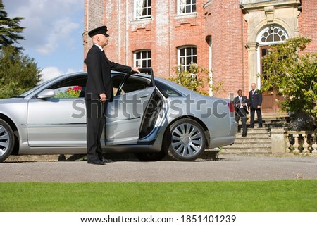 Chauffeur holding the car door open for two senior businessmen in the background in front of a huge manor house