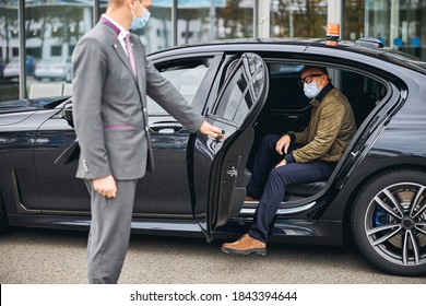 Chauffeur Opening The Car Door For A Caucasian Male Passenger In A Disposable Face Mask