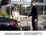 Chauffeur packs a suitcase in a car trunk, businesswoman waiting nearby using luxury taxi service during a business trip. Concept of business transfer services, idea of personal driver.
