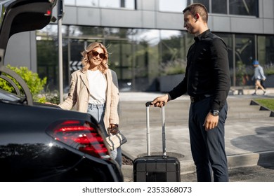 Chauffeur packs a suitcase in a car trunk, businesswoman waiting nearby using luxury taxi service during a business trip. Concept of business transfer services, idea of personal driver.