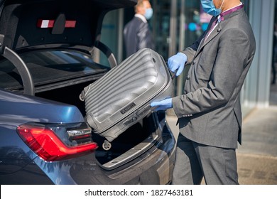Chauffeur in suit is packing luggage into car near airport terminal while wearing protective mask and gloves - Shutterstock ID 1827463181