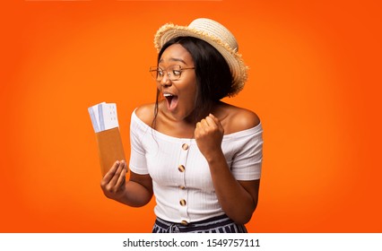 Cheap Flights. Excited Black Woman Traveler Holding Tickets And Passport Shaking Fists Standing On Orange Background. Studio Shot