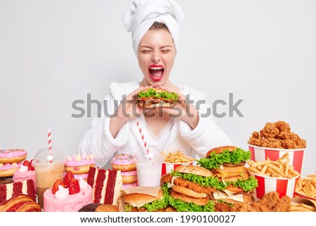 Cheat mea and gluttony. Funny young woman exclaims loudly keeps mouth wide opened eats tasty burger surrounded by variety of fast food wears bathrobe towel over head isolated over white background