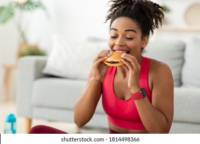 Cheat Meal. Black Fit Woman Eating Burger Enjoying Unhealthy Fast Food After Training Sitting On Floor At Home. Free Space