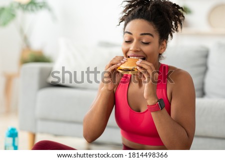 Cheat Meal. Black Fit Woman Eating Burger Enjoying Unhealthy Fast Food After Training Sitting On Floor At Home. Free Space