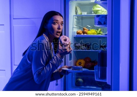 Cheat meal concept. Afraid lady eating donuts near opened fridge late in night, looking aside with scared face expression