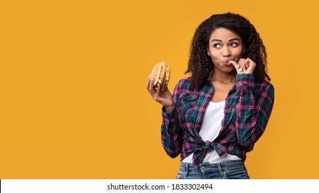 Cheat Meal. Portrait Of Happy African American Casual Woman Eating Tasty Burger, Licking Finger, Looking Away At Copy Space. Satisfied Woman Holding Fastfood, Isolated Over Orange Background, Banner - Shutterstock ID 1833302494