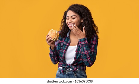 Cheat Meal. Portrait Of Happy African American Lady Eating Tasty Burger, Laughing And Wiping Lips With Finger, Copy Space. Satisfied Woman Holding Fastfood, Isolated Over Orange Background, Banner