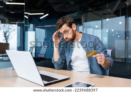 Cheated and disappointed businessman working inside office, man holding bank credit card and trying to make a purchase in online store, online fraud and deception.