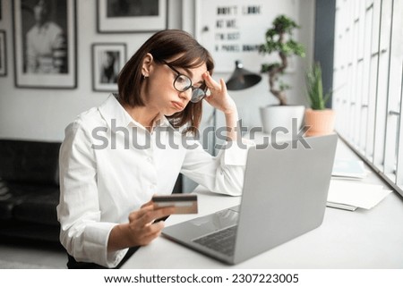 Cheated and disappointed young businesswoman working in cozy office or cafe, holding bank credit card and trying to make purchase, online fraud and deception