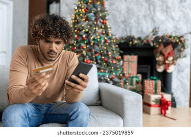 Cheated and upset Christmas man in living room near decorated Christmas tree, rejected and wrong money transfer, hispanic man holding bank credit card and phone.