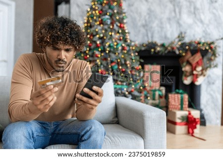 Cheated and upset Christmas man in living room near decorated Christmas tree, rejected and wrong money transfer, hispanic man holding bank credit card and phone.