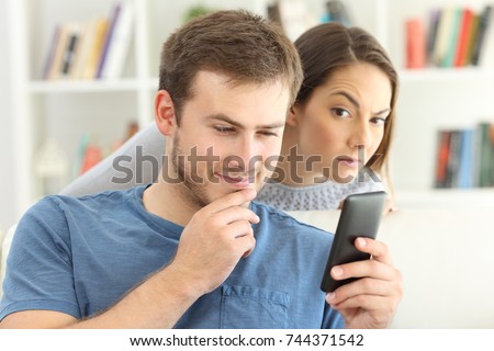 Cheater man dating on line with a smart phone and girlfriend is spying sitting on a sofa at home