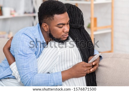 Cheating husband. Young african american man texting on cellphone with somebody while embracing his wife at home, closeup