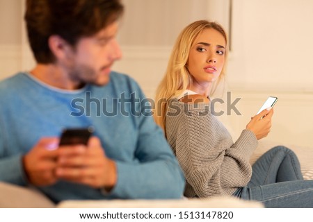 Cheating And Infidelity. Husband Texting On Cellphone Sitting Next To Suspicious Wife On Couch At Home. Selective Focus