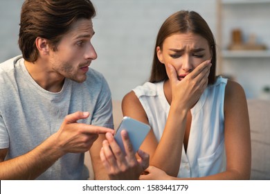 Cheating And Infidelity. Jealous Boyfriend Showing Phone To Crying Girlfriend Demanding Explanation Sitting On Couch Indoor. Selective Focus