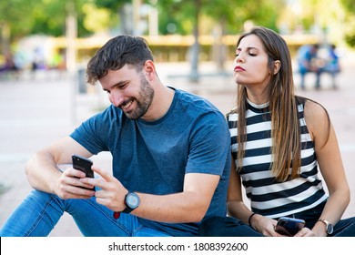 Cheating man chatting with women on his smartphone while his girlfriend spies on him sitting on a park bench - Shutterstock ID 1808392600