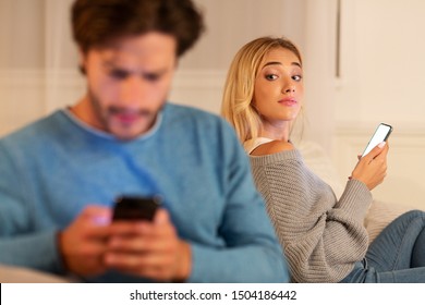 Cheating Signs. Girl Looking At Her Boyfriend Texting On Phone Suspecting Infidelity Sitting On Couch At Home. Selective Focus