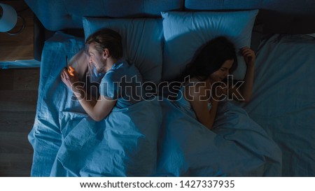 Cheating and Unfaithful Young Man Uses Smarphone for Finding a New Date or Bragging Night in Bed when Girlfriend Sleeps Soundly. Top View Camera