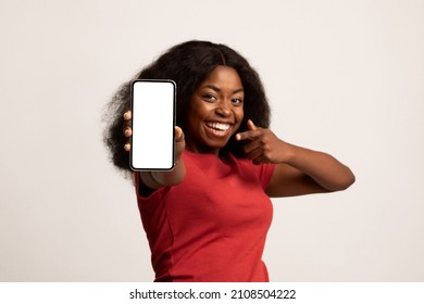 Check This Ad. Cheerful Black Woman Holding And Pointing At Blank Smartphone With White Screen, Happy Millennial African American Lady Recommending New App Or Mobile Website, Mockup Image