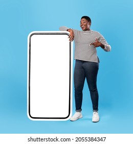 Check This Application. Black Female Showing Big Phone With Empty Screen Standing And Posing Near Huge Full Length Smartphone Over Blue Studio Background. Great App For Cellphone. Mockup, Square