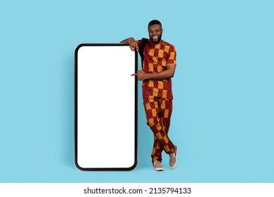 Check This. Cheerful Black Man In Traditional Costume Pointing At Big Blank Smartphone While Standing Over Blue Studio Background, Africa Male Showing Mobile Phone With White Screen For Mockup
