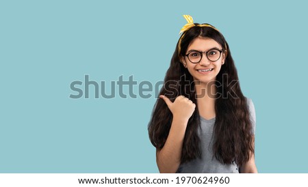 Check this. Cheerful Indian teen girl pointing aside with thumb up at empty space on blue studio background, banner design. Joyful adolescent advertising or promoting something, panorama