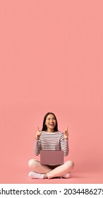 Check This. Excited Asian Female Sitting With Laptop And Pointing Up At Copy Space Above Her Head On Pink Studio Background, Cheerful Young Korean Woman Showing Free Place For Your Text Or Design