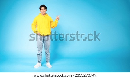 Check this. Happy young asian guy pointing up at free space for text, expressing positive emotion and demonstrating empty place for advertisement standing on blue background, full length, panorama