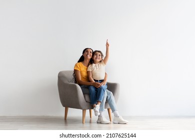 Check This. Happy Young Mother And Little Daughter Sitting In Armchair And Pointing Up, Cheerful Middle Eastern Mom And Female Child Demonstrating Copy Space Above Their Heads While Resting In Chair - Powered by Shutterstock