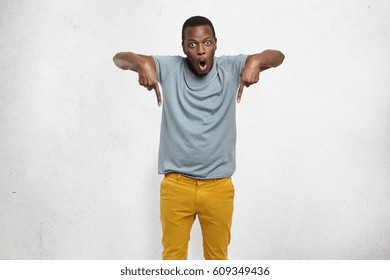 Check this out! Cropped studio shot of attractive excited young African male in t-shirt and mustard pants pointing fingers down having surprised look, his face expressing complete shock and disbelief - Powered by Shutterstock