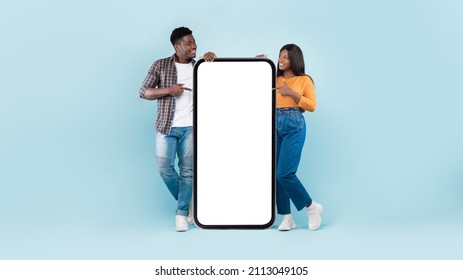 Check This. Portrait of cheerful young black couple leaning on white cell screen pointing fingers at empty display, recommending mobile app, mock up for website design advert, banner, blue studio wall