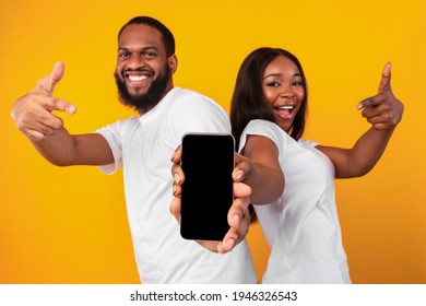 Check This. Positive African American man and woman holding and presenting mobile phone with black empty screen in hands, pointing fingers at gadget with blank space for mock up, yellow studio