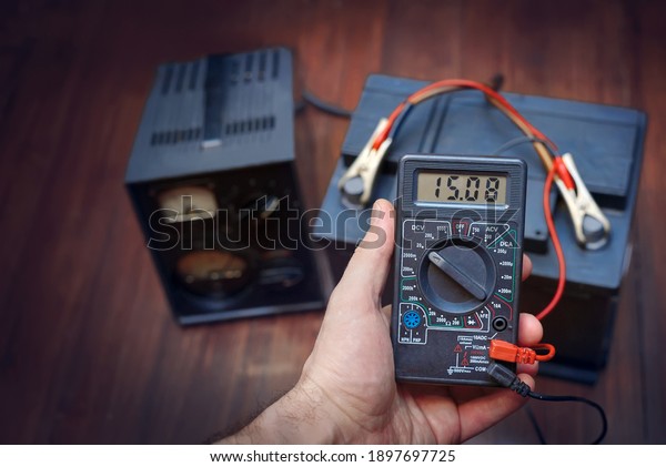 Checking and charging car battery with\
charger from home electricity. Man measures voltage. Mechanic using\
multimeter, voltmeter to check voltage level on car battery. Car\
battery maintenance