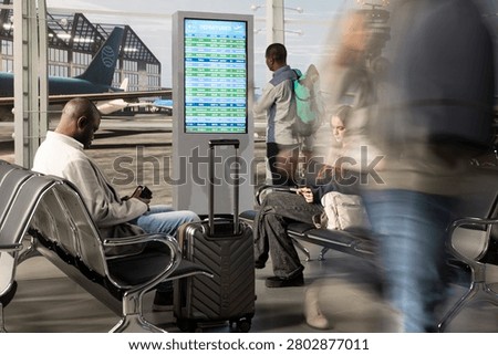 Checking electronic flights timetable, fast movement and multiethnic passengers sitting on airport benches, reading FIDS display schedule, high volume airport traffic and diverse group of people.