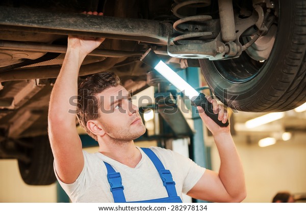 Checking\
every detail for defects. Confident young man in uniform holding\
lamp while standing underneath a car in\
workshop