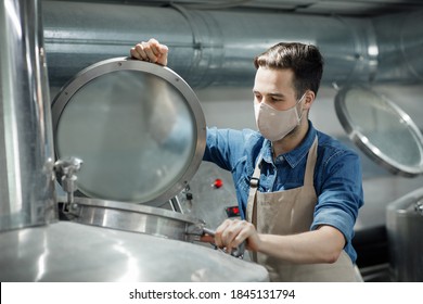 Checking of product in manufacturing factory during coronavirus pandemic. Attractive young male worker in apron and protective mask closes tank for starters fermentation of craft beer, free space - Powered by Shutterstock