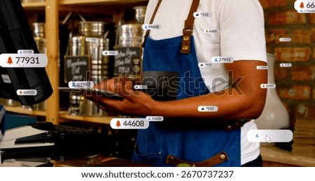 Checking tablet, barista in blue apron reviewing POS monitor behind wood counter in cafe, with vase. Hospitality, hospitalityindustry, retail, artisan, rustic, digital, customerservice