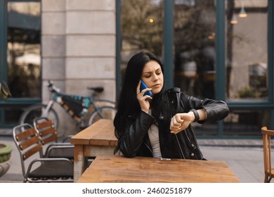 Checking time. Serious teen worried girl sitting in cafe or restaurant outside looking at her watch, have mobile phone conversation while waiting for her friends. Woman look angry or mad.