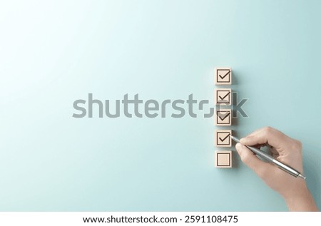 Checklist completion and task management concept. A hand holding a pen marks checkboxes on wooden blocks arranged in a vertical list, symbolizing organization, task management, and goal achievement.
