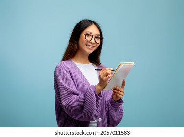 Checklist concept. Asian female student in glasses, taking notes in copybook and smiling to camera on blue studio background. Korean lady studying, preparing for exam, writing in notebook