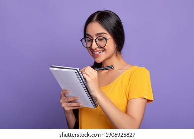 Checklist. Portrait of intelligent indian female student wearing glasses, taking notes in copybook on purple studio background. Smart young woman studying, preparing for exam, writing in notebook